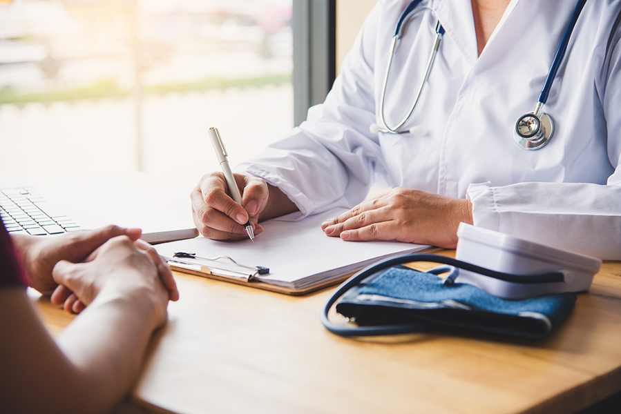 Ein Arzt sitzt am Schreibtisch und macht Notizen. Ein Patient sitz gegenüber. Das Bild ist so gezoomt, dass die Personen nicht zu erkennen sind. Ein Arzt sitzt am Schreibtisch und macht Notizen. Ein Patient sitz gegenüber. Das Bild ist so gezoomt, dass die Personen nicht zu erkennen sind.